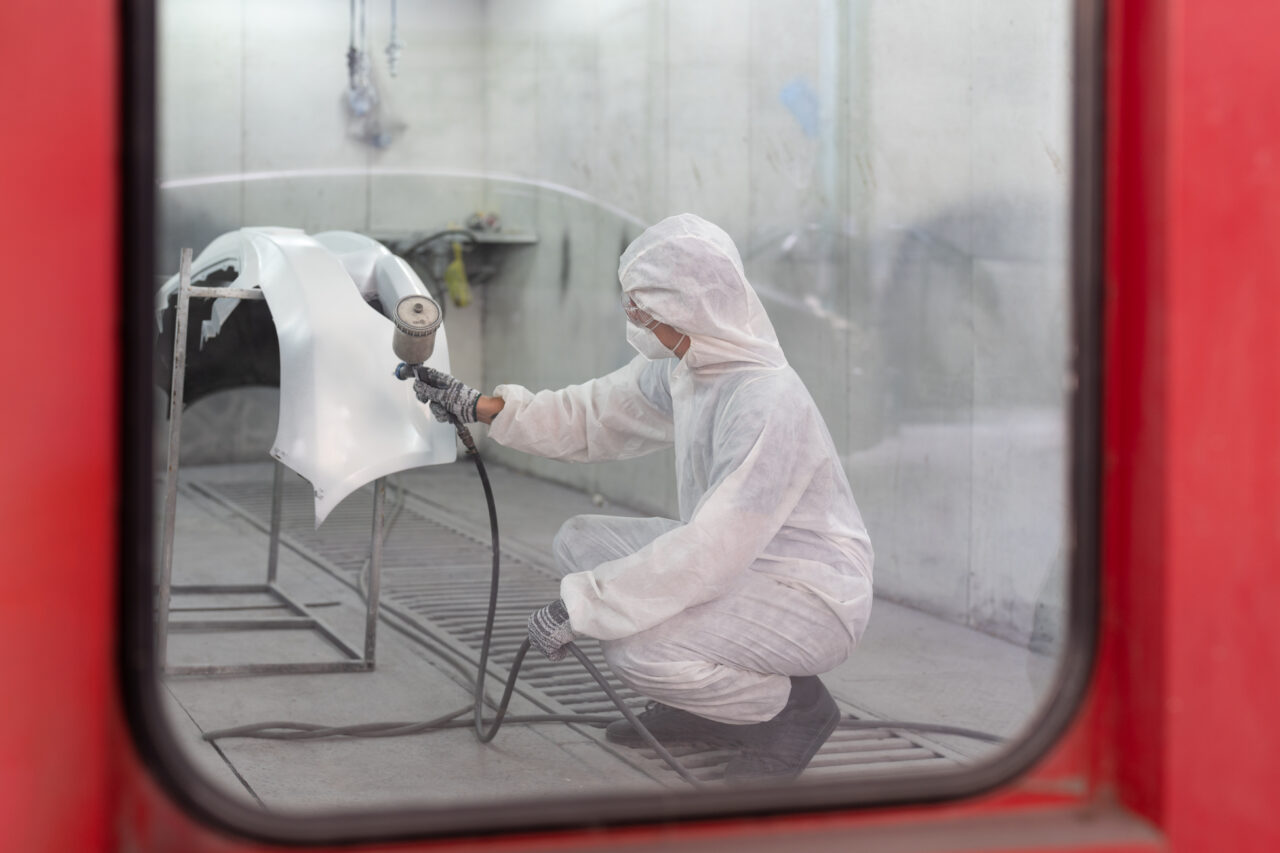 Automobile repairman painter in protective workwear painting part of car in paint chamber at auto car garage service and paint center.